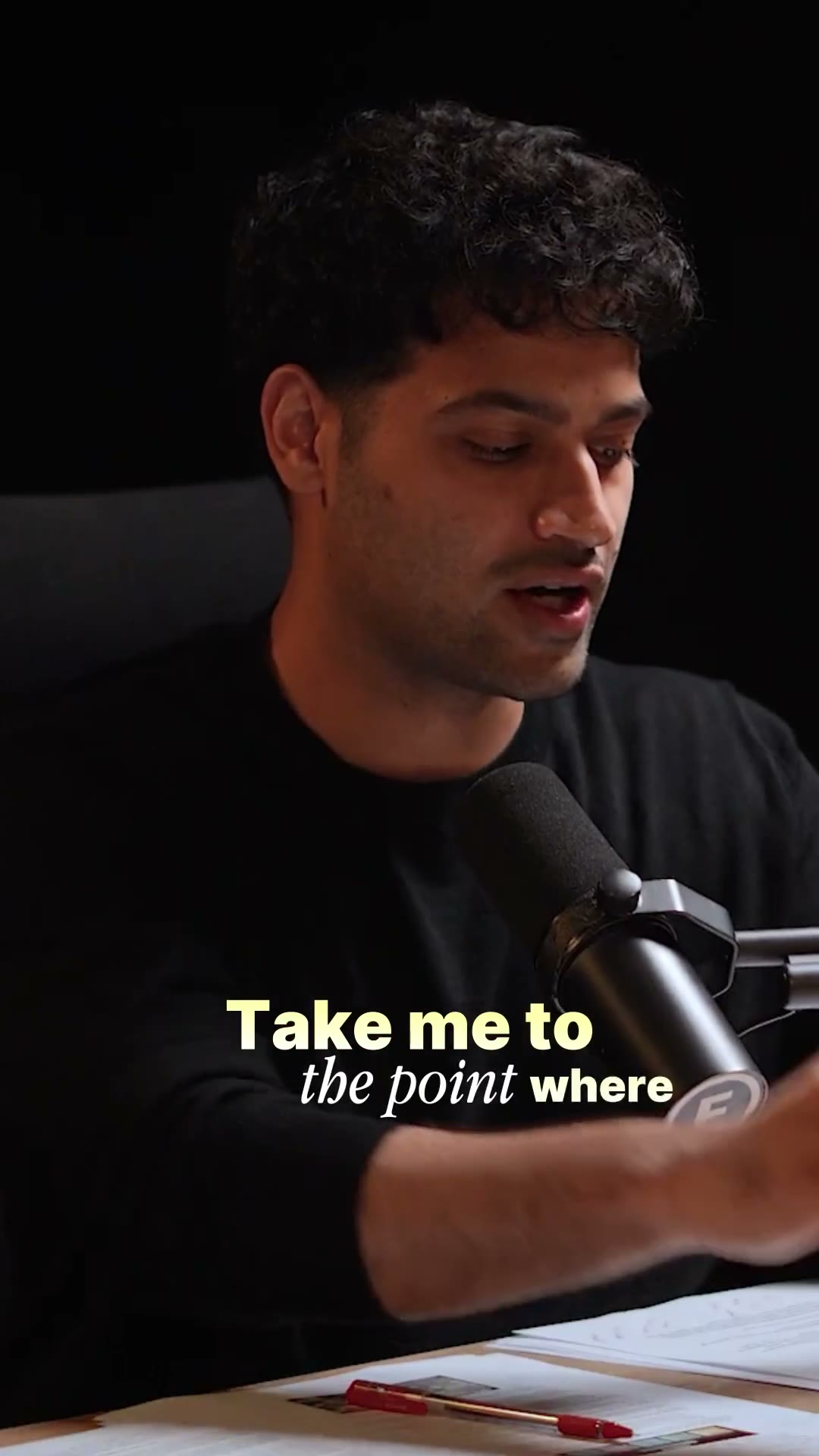 A man in a black tee leans into a boom-mounted studio mic against a black set, a yellow-and-white burned-in caption reading “Take me to the point where” across his chest.